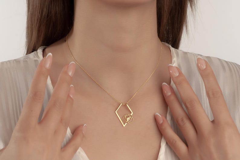 May include: A gold-colored necklace featuring a diamond-shaped pendant with a floral design at the center. The necklace is suspended from a delicate chain. The image shows the necklace resting on a person's chest.