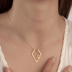 May include: A gold-colored necklace with a geometric pendant. The pendant features a diamond-shaped frame with a flower design inside. The necklace is worn on a person with a white top.