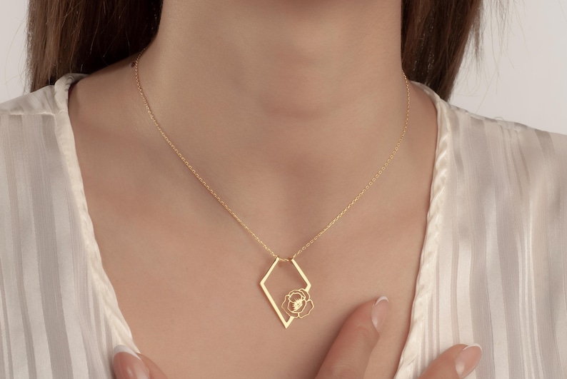 May include: A gold-colored necklace with a geometric pendant. The pendant features a diamond-shaped frame with a flower design inside. The necklace is worn on a person with a white top.