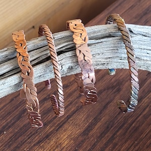 May include: Four copper bracelets with unique designs. Two have a braided pattern, one has a twisted rope design, and the last has a twisted design with silver accents. The bracelets are displayed on a piece of driftwood.
