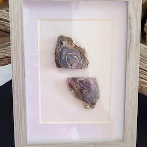 May include: A light gray wood-grain frame containing a white mat board with two polished agate slices mounted vertically. The top agate slice features swirls of purple, gray, and white, while the bottom slice displays shades of purple and brown.