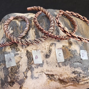 May include: Four copper-colored metal cuff bracelets, each featuring a twisted rope design. The bracelets are arranged in a row and labeled with small paper tags marked with the letters A, B, C, and D, indicating four distinct options.