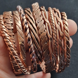 May include: A collection of copper-colored bracelets with various braided and textured designs. The bracelets are displayed in a hand, showcasing the different styles and finishes. The warm tones of the metal create a visually appealing display of jewelry.