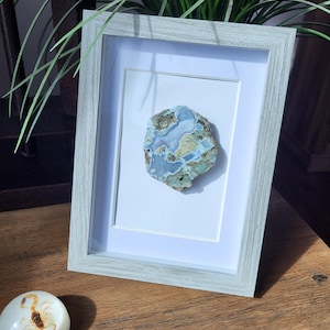 May include: A framed, polished stone with blue, green, and brown hues. The stone is mounted on a white mount within a light grey wooden frame. A decorative, white, oval-shaped object with a scorpion design is in the foreground.