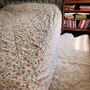 May include: A quilted bedspread featuring a repeating floral pattern in shades of orange, yellow, and green on a white background, draped over a bed with a black metal frame, positioned next to a wooden nightstand filled with various books.