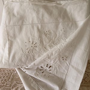 May include: A white fabric textile featuring intricate cutwork embroidery of floral patterns and leaves. The piece has a hemmed border with decorative stitching and a smooth, lightweight texture throughout the folded material.