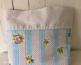 Puede incluir: Un bolso de tela rectangular que presenta una sección superior con fondo blanco y lunares negros. La sección inferior muestra rayas verticales de color azul claro, lunares negros y pequeños ramos florales en rosa, amarillo y azul.
