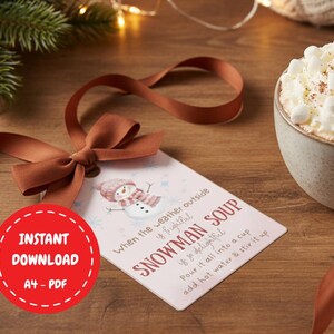 Könnte beinhalten: Ein Geschenk-Anhänger mit einem Schneemann-Motiv und dem Schriftzug "Snowman Soup". Der Anhänger ist mit einem braunen Band gebunden. Ein roter Kreis mit "Instant Download A4-PDF" ist ebenfalls sichtbar.