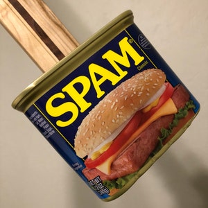 May include: A can of Spam, with the label showing a Spam sandwich with cheese, tomato, lettuce, and mustard on a sesame seed bun. The can is gold and blue with the word "SPAM" in yellow letters. The label also says "Classic" and "NET WT 12 OZ (340g)".