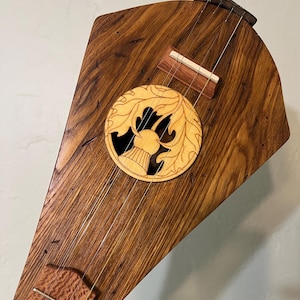 May include: A handcrafted wooden dulcimer with a natural wood grain finish. The instrument features a decorative circular inlay with a leaf and antler design. Strings are stretched across the body, and a bridge is visible. The dulcimer is a folk music instrument.