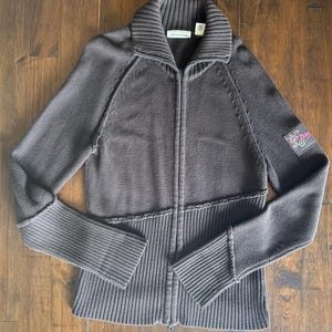 May include: A dark brown zip-up jumper featuring a ribbed collar, long sleeves, and a full-length front zip. The garment includes a textured knit pattern on the upper torso and sleeves, with a patch on the left arm displaying the text DKNY.