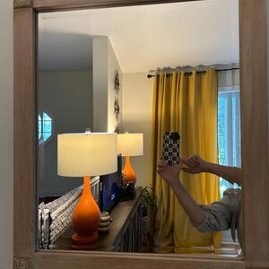 May include: A rectangular wall mirror featuring a light-toned, natural wood frame with decorative square corner blocks. The mirror reflects a room interior containing orange table lamps, yellow curtains, and a piece of furniture.