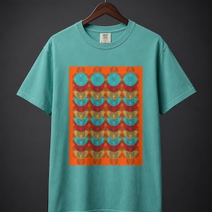 Könnte beinhalten: Ein kurzärmeliges T-Shirt in Blaugrün mit einem rechteckigen Grafikdruck auf der Brust. Das Motiv besteht aus einem sich wiederholenden Muster aus überlappenden Kreisen und Halbkreisen in Blau-, Rot- und Goldtönen auf leuchtend orangefarbenem Grund.