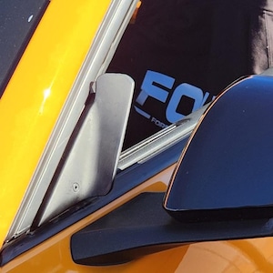 May include: Close-up of a black side mirror on a yellow car. The mirror is sleek and modern, reflecting the surroundings. The car's body is a vibrant yellow, with a black trim. The image captures the details of the car's exterior.