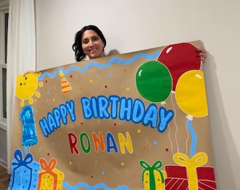 Può includere: Un grande striscione di compleanno rettangolare in carta marrone con la scritta "HAPPY BIRTHDAY RONAN" in lettere blu e multicolori. Il design include illustrazioni dipinte di un palloncino blu a forma di numero uno e pacchi regalo.
