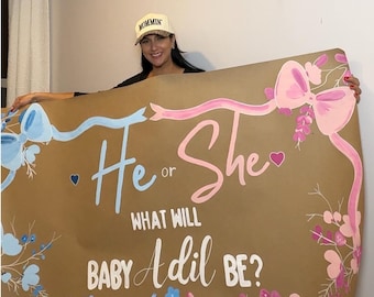May include: A large rectangular brown paper sign featuring hand-painted blue and pink floral accents, ribbons, and bows. The text reads: He or She, What Will Baby Adil Be? The sign includes small blue and pink hearts and decorative botanical illustrations.