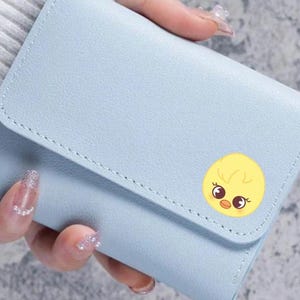 Könnte beinhalten: Hellblaues Portemonnaie mit Klappenverschluss. Das Portemonnaie hat einen gelben Kükenaufkleber auf der Vorderseite. Das Portemonnaie wird von einer Person gehalten.
