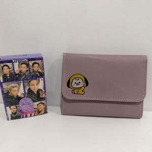 May include: A light purple wallet with a cartoon character design and a small purple box with photos of people on it. The wallet has a flap closure and a stitched border. The box has the word "BTS" on it.