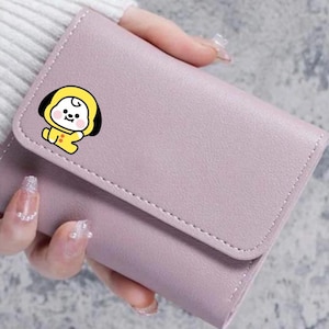 Könnte beinhalten: Ein helllila, dreifach gefaltetes Portemonnaie mit einer genähten Kante. Das Portemonnaie hat eine gelbe Cartoonfigur mit schwarzem Umriss auf der Vorderseite. Das Portemonnaie wird in einer Hand gehalten.