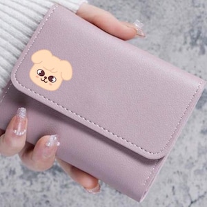 Könnte beinhalten: Ein lila-farbenes, zweifach gefaltetes Portemonnaie mit einer genähten Bordüre und einem Klappenverschluss. Das Portemonnaie hat ein Cartoon-Hunde-Gesicht auf der Vorderseite. Das Portemonnaie wird in einer Hand gehalten.