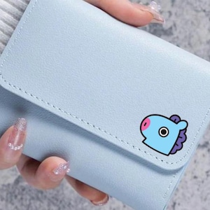 Könnte beinhalten: Hellblaues Portemonnaie mit Klappe. Es hat eine Cartoonfigur mit blauem Kopf, rosa Nase und lila Mähne. Das Portemonnaie ist aus glattem Material und hat einen genähten Rand.