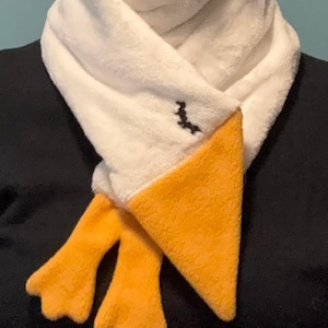May include: A white scarf with a duck design. The scarf has a white body, a yellow beak, and yellow feet. A small black embroidered design is on the scarf. The scarf is made of a soft, plush material.