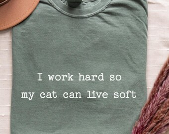 Puede incluir: Camiseta verde salvia con el texto "I work hard so my cat can live soft" en blanco. Un sombrero marrón y un ramo de flores rosas secas también están en la imagen. La camiseta está doblada sobre una superficie blanca.