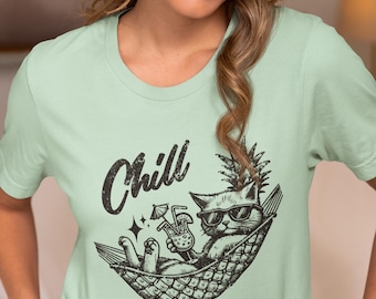 May include: Light green t-shirt with a graphic of a cat in sunglasses relaxing in a hammock, holding a cocktail. The word "Chill" is above the cat, and the text "WORK HARD SO YOUR CAT CAN LIVE SOFT" is below.