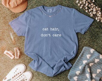 Pode incluir: Uma camiseta azul com o texto branco "cat hair, don't care". A camiseta é exibida com um colar, chapéu, sapatos e grampos de cabelo. A camiseta é feita de material macio.