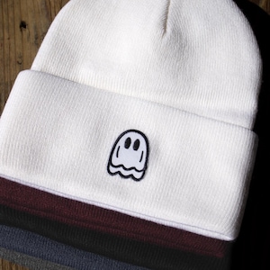 May include: A stack of four cuffed knit beanies in white, burgundy, black, and grey. The top white beanie features a small, centered embroidered patch of a white ghost with a black outline and two black oval eyes on the folded cuff.