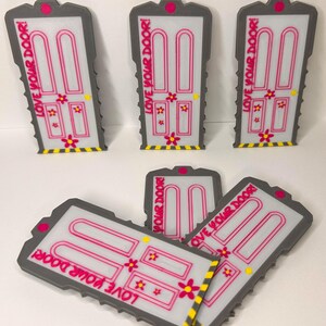 May include: A collection of six gray, white, and pink door-shaped ornaments. Each ornament features the text LOVE YOUR DOOR! in pink, with pink flower accents, a yellow doorknob, and a yellow and black striped pattern along the bottom edge.