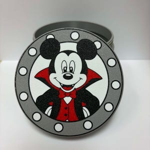 May include: A round metal container lid featuring a graphic of a cartoon mouse dressed as a vampire with a black cape, red vest, and bow tie. The design is framed by a grey circular border with white dots, set against a white background.