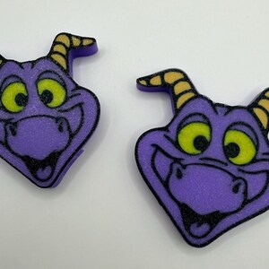 May include: Two purple dragon head charms with yellow horns and bright green eyes. Each charm features a smiling expression with black outlines and a textured surface. The dragon heads are shaped like a diamond and have small horns on top.