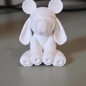 May include: A white 3D-printed figurine of a seated animal with large ears and a rounded head. The figurine has a smooth texture and is sitting on a gray surface. The ears are round, and the body is in a sitting position.