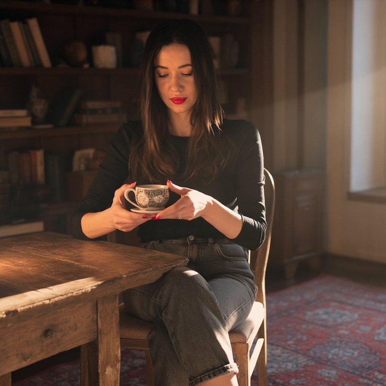 Puede incluir: Una persona sentada en una mesa de madera, sosteniendo una peque&ntilde;a taza de t&eacute; y platillo. La taza tiene un dise&ntilde;o decorativo. Lleva una camisa negra de manga larga y vaqueros. El fondo incluye una estanter&iacute;a y una alfombra estampada.