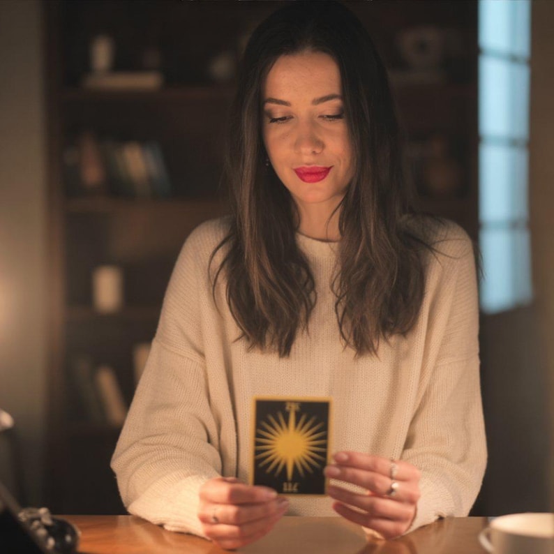Puede incluir: Una persona sostiene una carta de tarot con un dise&ntilde;o de sol amarillo sobre un fondo negro. La carta tiene el n&uacute;mero 11C en la parte inferior. La persona lleva un su&eacute;ter color crema y anillos.