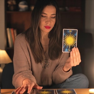 Puede incluir: Una mujer sostiene una carta del tarot con un dise&ntilde;o de sol amarillo sobre un fondo azul. La carta est&aacute; etiquetada como "IX". Lleva un su&eacute;ter beige y tiene pintalabios rojo. Hay otras cartas del tarot sobre la mesa frente a ella.