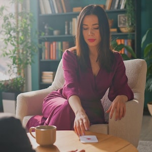 Puede incluir: Una mujer con un vestido color ciruela est&aacute; sentada en un sill&oacute;n crema, sosteniendo una carta con un coraz&oacute;n rojo sobre una mesa de madera. Una taza marr&oacute;n est&aacute; sobre la mesa. Una estanter&iacute;a y plantas est&aacute;n en el fondo.
