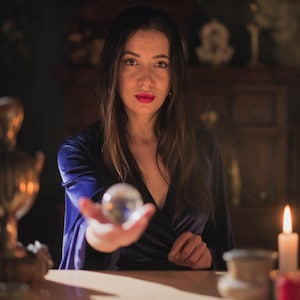 Puede incluir: Una mujer con cabello casta&ntilde;o largo y l&aacute;piz labial rojo sostiene una bola de cristal transparente. Lleva un vestido de terciopelo azul oscuro. La escena est&aacute; tenuemente iluminada con velas y decoraci&oacute;n antigua.