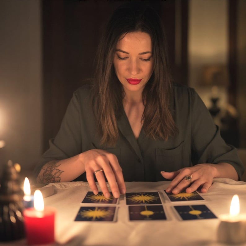 Puede incluir: Una persona est&aacute; sentada a una mesa, colocando cartas del tarot. Las cartas son rectangulares con un dise&ntilde;o de sol. Velas encendidas en rojo y blanco iluminan la escena, creando un ambiente c&aacute;lido e &iacute;ntimo. La persona lleva una camisa verde y tiene el pelo largo y casta&ntilde;o.