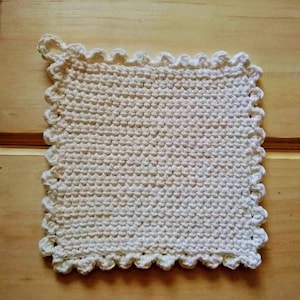 May include: A square, off-white crochet potholder with a scalloped edge and a loop for hanging. The handmade kitchen item is displayed on a light wood surface. The crochet stitch creates a textured appearance.