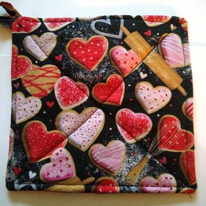 May include: Square quilted pot holder with a black background and a pattern of heart-shaped cookies in red, pink, and white. A rolling pin and whisk are also featured in the design. A loop is attached for hanging.