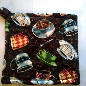 May include: A square quilted fabric potholder featuring a dark brown background with a repeating pattern of various coffee cups, saucers, and spoons. The design includes cups in shades of blue, green, and red with checkered or polka dot patterns.