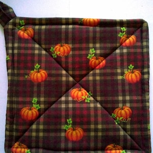 May include: Square quilted potholder with a plaid pattern in shades of brown, red, and green. The potholder is decorated with orange pumpkins and green vines. It has a loop for hanging.