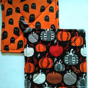 May include: Two Halloween-themed pot holders. One is orange with black ghost shapes. The other is black with orange, gray, and white pumpkin designs. Both have quilted stitching and a loop for hanging.