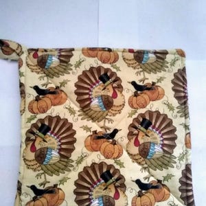 May include: A beige quilted pot holder with a Thanksgiving theme. The pot holder features a repeating pattern of turkeys wearing top hats and pumpkins.
