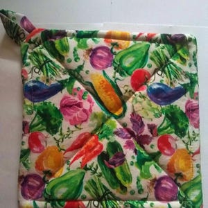 May include: Square quilted pot holder with a loop for hanging. The fabric features a colorful pattern of various vegetables, including corn, tomatoes, peppers, and eggplant, on a white background. The edges are neatly sewn.