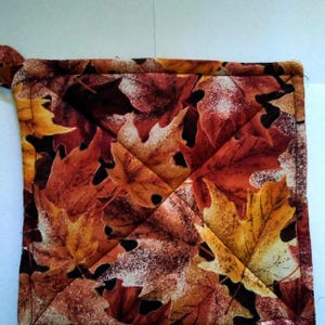 May include: A square quilted pot holder featuring a fall leaf pattern in shades of orange, brown, and yellow. The pot holder has a loop for hanging and a stitched diamond pattern across the surface.