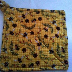 May include: A square quilted potholder featuring a yellow and orange gingham check background. The fabric is printed with a repeating pattern of bumblebees, brown pinecones, green pine needles, and small red floral accents throughout.