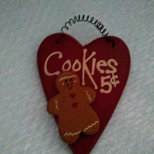 May include: A heart-shaped red wooden sign featuring a brown gingerbread man figure and the hand-painted text "Cookies 5¢" in a light tan color. The sign includes a black coiled wire hanger attached to the top for wall mounting purposes.